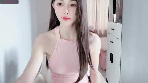 Media: Video of a young Asian woman with long, straight, dark brown hair, wearing a pink sleeveless top. She stands indoors near a white dresser with drawers and a pink blanket.