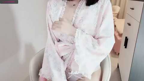 Media: Video of a fair-skinned person in a light pink, lace-trimmed robe, partially unzipped to reveal a small breast, seated in a beige chair against a neutral-colored backdrop.