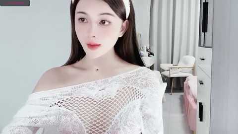 Media: Video of a young, pale-skinned woman with long dark hair, wearing a white lace off-shoulder top, standing in a modern, bright beauty salon with white furniture and makeup products.