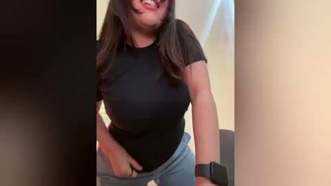 Media: Video of a smiling, light-skinned woman with long dark hair, wearing a black t-shirt and light blue jeans, standing indoors with a black smartwatch on her wrist.