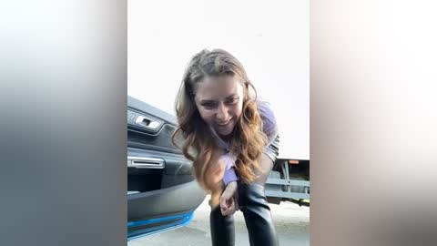 Media: Video of a young woman with long, wavy brown hair, bending over in a parking lot. She wears a purple long-sleeve shirt and black pants. A grey car is parked behind her.