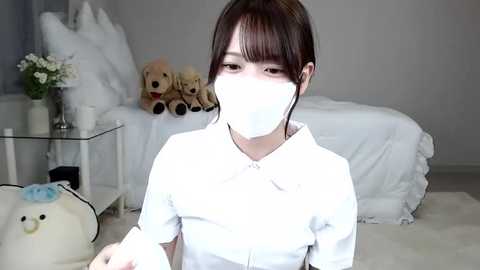 Media: Video of an Asian woman with straight dark hair, wearing a white mask, white top, and standing in a minimalist bedroom with white bedding, teddy bears, and a vase of flowers.