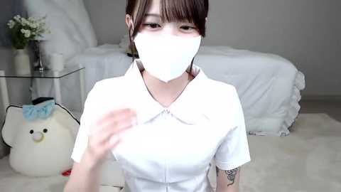 Media: Video of an East Asian woman with light skin, short brown hair, and bangs, wearing a white nurse outfit and mask, kneeling on a plush white carpet. Background includes a bed, vase, and stuffed bear.