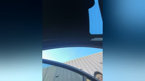 Media: A video showing a person's face inside a car, looking out the window at a blue sky and a grey corrugated metal roof. The image is slightly out of focus.