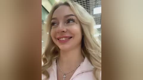 Media: Video of a smiling, fair-skinned woman with long, wavy blonde hair, wearing a pink zip-up hoodie and a silver cross necklace. She is standing in a brightly lit, modern indoor setting with a grid-patterned ceiling.