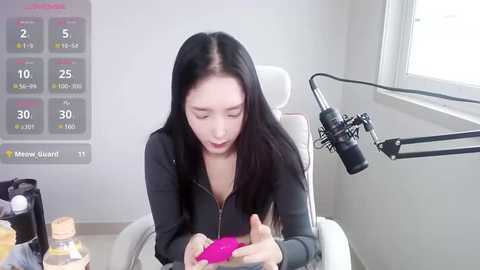 Media: A video of an East Asian woman with long black hair, wearing a black jacket, using a pink vibrator on a white chair. Background includes a white wall, a window, and a microphone stand.