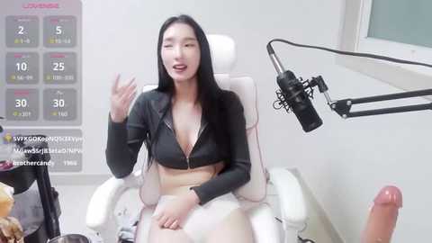 Media: A video of an East Asian woman with long black hair, wearing a black zip-up jacket and white shorts, seated on a white chair, with a microphone and a large erect penis on a table in the background.