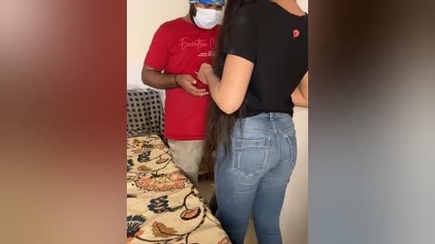 Media: A video shows a dark-skinned man in a red shirt and beige pants holding a light-skinned woman with long hair in a black shirt and tight jeans, standing in a laundry room with a patterned bedspread.