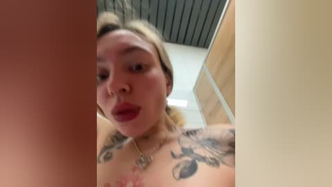 Media: Video of a woman with light skin, blonde hair, and tattoos on her chest, wearing red lipstick, looking surprised. Background shows a beige wall and a wooden door.