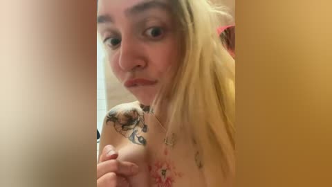 Media: A close-up video of a young woman with pale skin and long blonde hair, wearing a choker, with visible tattoos on her chest and neck, captured in a bathroom with beige tiles and a red towel.