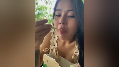 Media: A video of a young Asian woman with long black hair, wearing a floral bikini top, eating a slice of cake. She's indoors with blurred tropical foliage in the background.