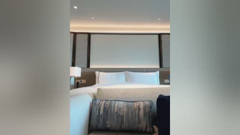 Media: A video of a modern, minimalist hotel room with a plush, textured headboard, white bedding, and a framed window with vertical blinds.