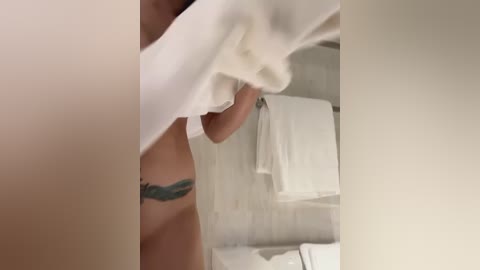 Media: Video of a person with light skin and a tattoo of a teal feather on their left side, partially covered by a white towel, in a bathroom with white tiled walls and a towel hanging on a rack.