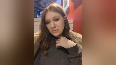 Media: Video of a young woman with long brown hair, wearing a dark sweater, sitting in a cozy room with red walls and a beige blanket.