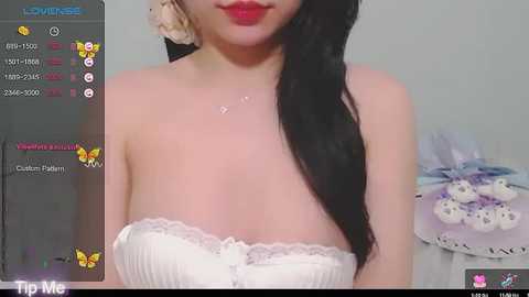 Media: Video of an Asian woman with fair skin, long black hair, wearing a white strapless lace dress. Background includes a live stream overlay with chat messages and virtual butterflies.