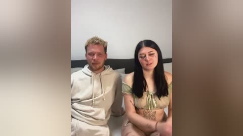 Media: Video of a pale-skinned man with curly blonde hair in a beige hoodie, and a pale-skinned woman with long black hair in a green off-shoulder top, sitting side by side on a couch.