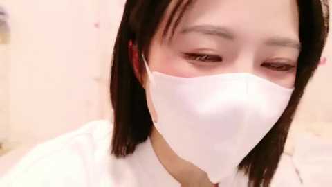 Media: Video of an Asian woman with medium skin tone, straight black hair, wearing a white surgical mask, looking downward with a neutral expression. Background is blurred, featuring soft pink walls.