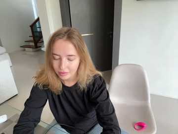 Media: Video of a young Caucasian woman with blonde hair, wearing a black sweater, sitting on a white chair in a minimalist, modern living room with a staircase and white walls.