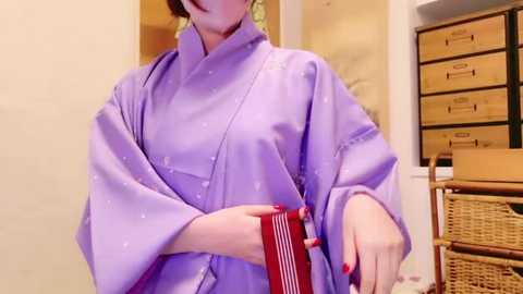 Media: Video of a person wearing a light purple, wide-sleeved kimono with a high collar, holding a red and white striped towel. Background features beige walls, a wooden dresser, and woven baskets.