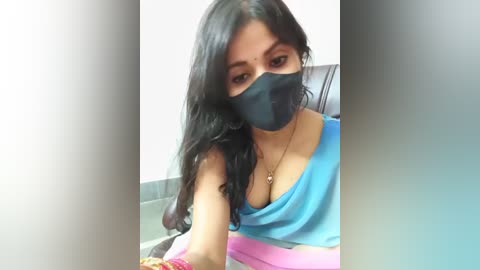 Media: Video of a South Asian woman with long black hair, wearing a blue tank top, black face mask, and pink bracelet, sitting indoors.