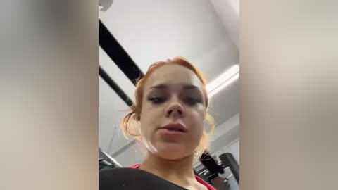 Media: Video of a young woman with light skin and red hair, wearing a black shirt, standing in a gym with a black exercise machine in the background.