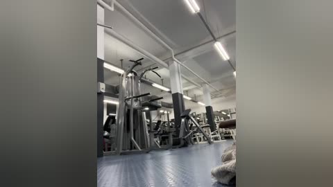 Media: Video of a modern, well-lit gym with a clean, white and grey color scheme. Visible equipment includes weight benches, dumbbells, and machines. A person in a black shirt is working out on the left.