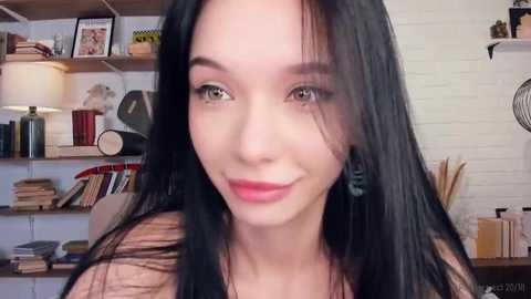 Media: A video of a young Asian woman with long black hair, fair skin, and brown eyes, wearing red lipstick and a black top. Background shows a white brick wall, shelves with books, and a lamp.