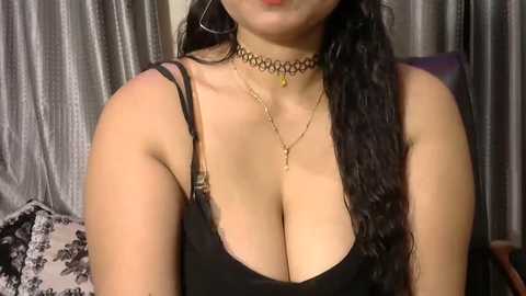 Media: Video of a light-skinned woman with long, wavy black hair, wearing a black spaghetti-strap top that reveals cleavage, a black choker, and gold necklace. Background shows grey curtains and a black floral-patterned cushion.