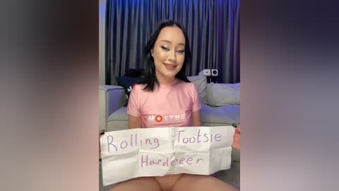 Media: Video of a smiling Asian woman with black hair, wearing a pink t-shirt, holding a sign that reads \"Rolling, Toesie, Hardcore.\" Background shows a living room with grey curtains and a couch.