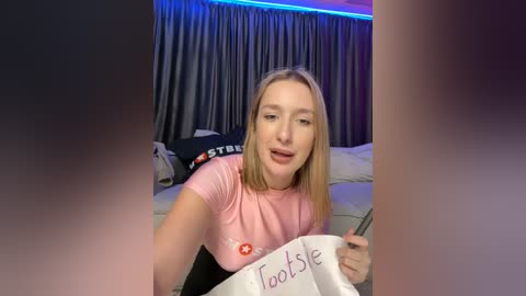 Media: Video of a young woman with blonde hair, wearing a pink shirt, holding a sign that reads \"Tadase,\" in a dimly lit room with grey curtains and a bed in the background.