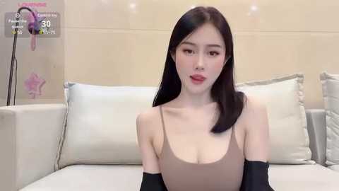 Media: A video of an East Asian woman with long black hair, fair skin, and red lipstick, wearing a beige spaghetti-strap top, sitting on a white couch. The background shows a minimalist room with beige walls and a pink plant.