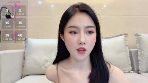 Media: A video of an East Asian woman with long black hair and fair skin, wearing a beige spaghetti-strap top, sitting on a light-colored sofa.