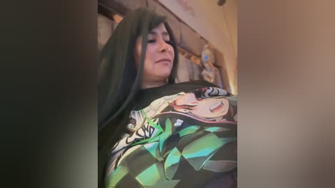 Media: Video of a person with long black hair, wearing a colorful anime T-shirt, standing in a dimly lit room with a plush toy in the background.