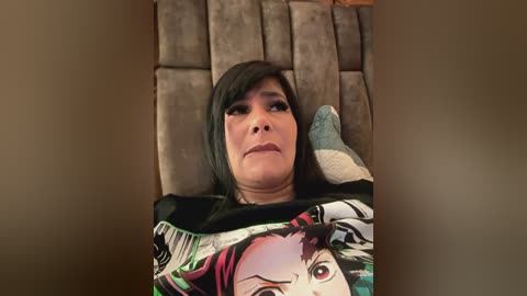 Media: Video of a middle-aged woman with dark hair, wearing a black T-shirt with a colorful anime character design, reclining on a beige, tufted sofa.