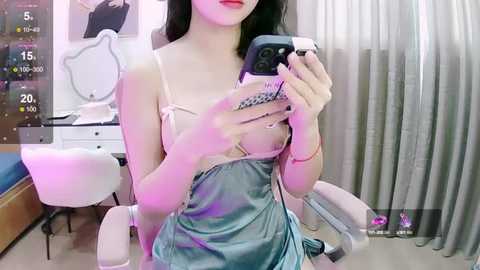 Media: Video of a young Asian woman with long black hair, wearing a revealing blue satin dress, taking a mirror selfie in a modern bedroom with a vanity table.