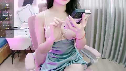 Media: Video of a young woman with long black hair, light skin, and a small bust, wearing a light blue satin slip dress, taking a selfie in a bedroom with white furniture, curtains, and a chair.