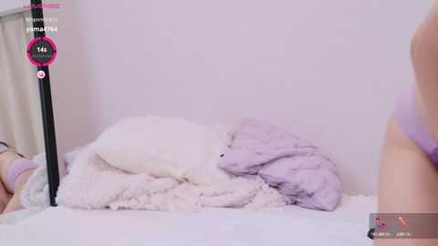 Media: Video of a soft, pastel-colored bedroom with a bed covered in white and lavender blankets, featuring a black bedside table and a pink and white digital overlay displaying a countdown.