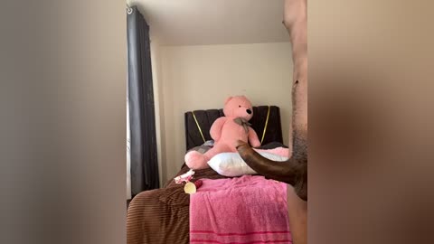 Media: Video of a man with dark skin, shirtless, holding a pink teddy bear on a bed with a pink blanket. The room has beige walls, dark curtains, and a simple, uncluttered appearance.