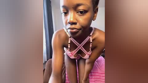 Media: Video of a young Black girl with short hair, wearing a pink bra with crisscross straps, leaning forward on a brown couch.