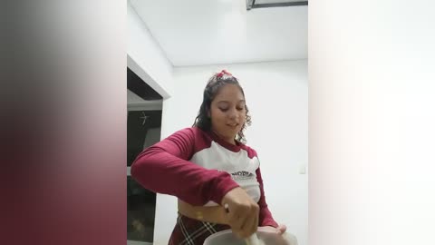 Media: Video of a Latina woman with curly hair in a red sweater and plaid skirt, cooking in a modern kitchen with white walls and a black ceiling light.