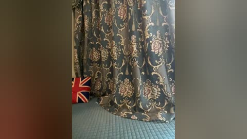 Media: Video of a room with a dark blue, ornate, floral-patterned curtain covering a window, revealing a Union Jack flag on the floor. The carpet is a textured, light blue.