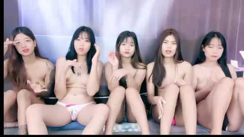 Media: Video of five East Asian women, topless, sitting on a bed, diverse in breast size and skin tone, some wearing pink panties. Background features soft, purple-hued curtains.