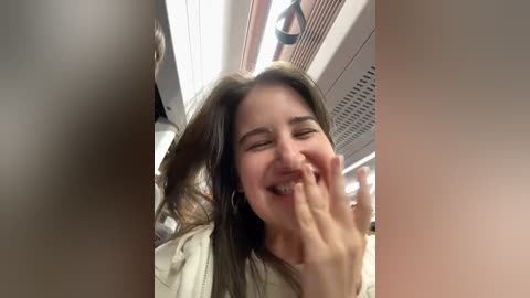 Media: A candid video captures a young woman with long brown hair, smiling and covering her mouth with her hand in a public elevator. She wears a white jacket. The background shows the elevator's metallic interior.