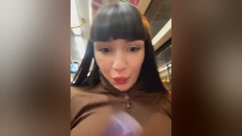 Media: A video of a young woman with long, straight black hair and blunt bangs, wearing a brown jacket, puckering her lips, taken from a blurred, close-up perspective.