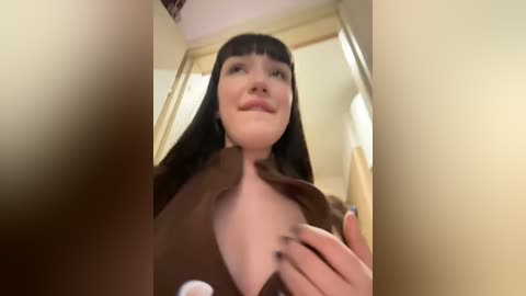 Media: Video of a young woman with long, straight black hair and pale skin, wearing a brown jacket, slightly lifting it to reveal cleavage. Background shows a beige wall and a mirror.