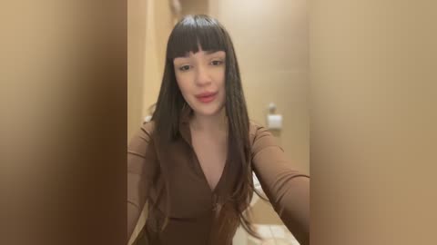 Media: Video of a young Asian woman with long, straight black hair, blunt bangs, and a V-neck, brown, long-sleeved top. She is standing in a narrow hallway with beige walls, looking slightly to the left.