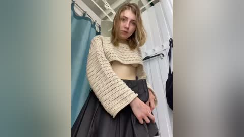 Media: Video of a young Caucasian woman with fair skin and shoulder-length blonde hair, wearing a cream knitted crop top and a pleated black skirt, standing in a modern bathroom with white tiles and a shower curtain.