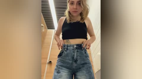 Media: Video of a young woman with shoulder-length blonde hair, wearing a black tank top and high-waisted, distressed blue jeans. She stands in a narrow, beige hallway with a wooden floor and a ceiling light strip.