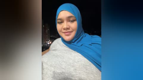 Media: Video of a smiling woman in a bright blue hijab and gray shirt, set against a dark, city night background.