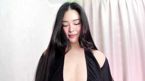 Media: Video of a young Asian woman with long, straight black hair, fair skin, wearing a black halter top, standing in a room with white and beige curtains in the background.